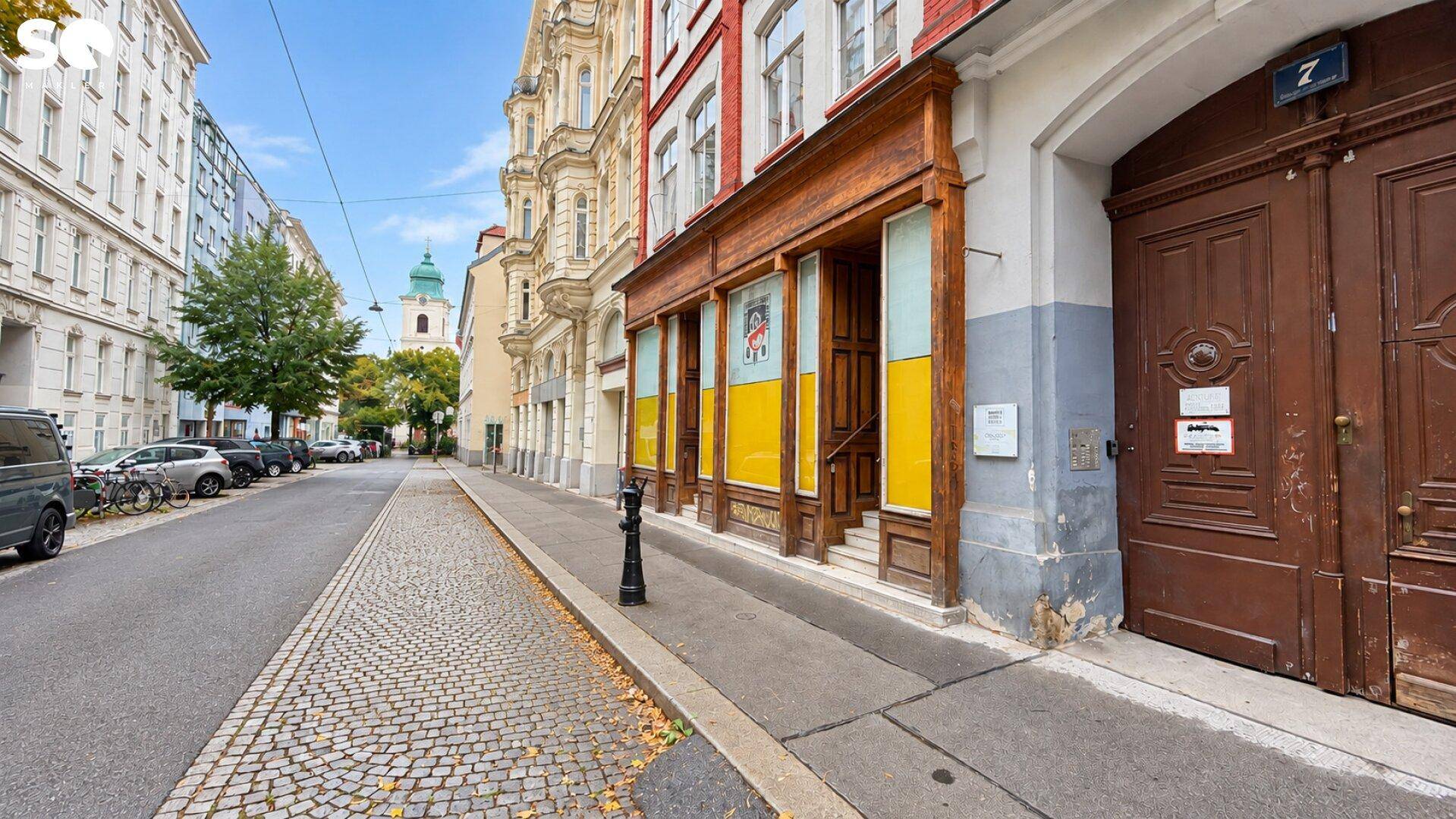 Historische Straßenansicht mit einem Gebäude, Geschäften im Erdgeschoss und einem Kirchturm im Hintergrund.