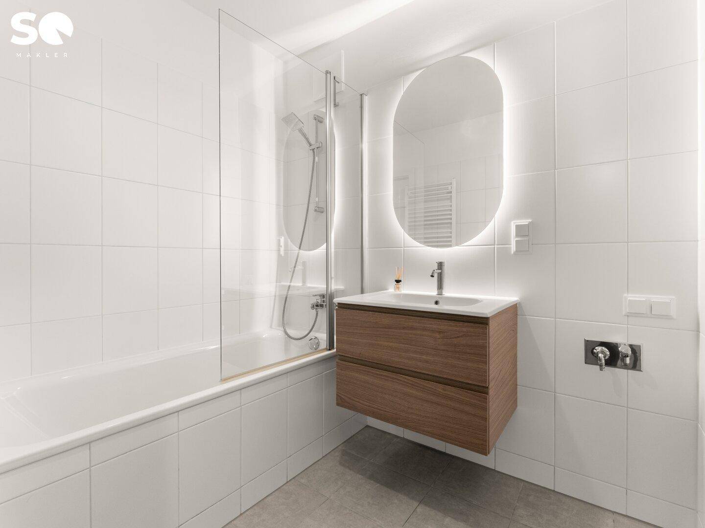 Modernes Badezimmer mit weißen Fliesen, Badewanne und einem beleuchteten Spiegel über dem Waschbecken.