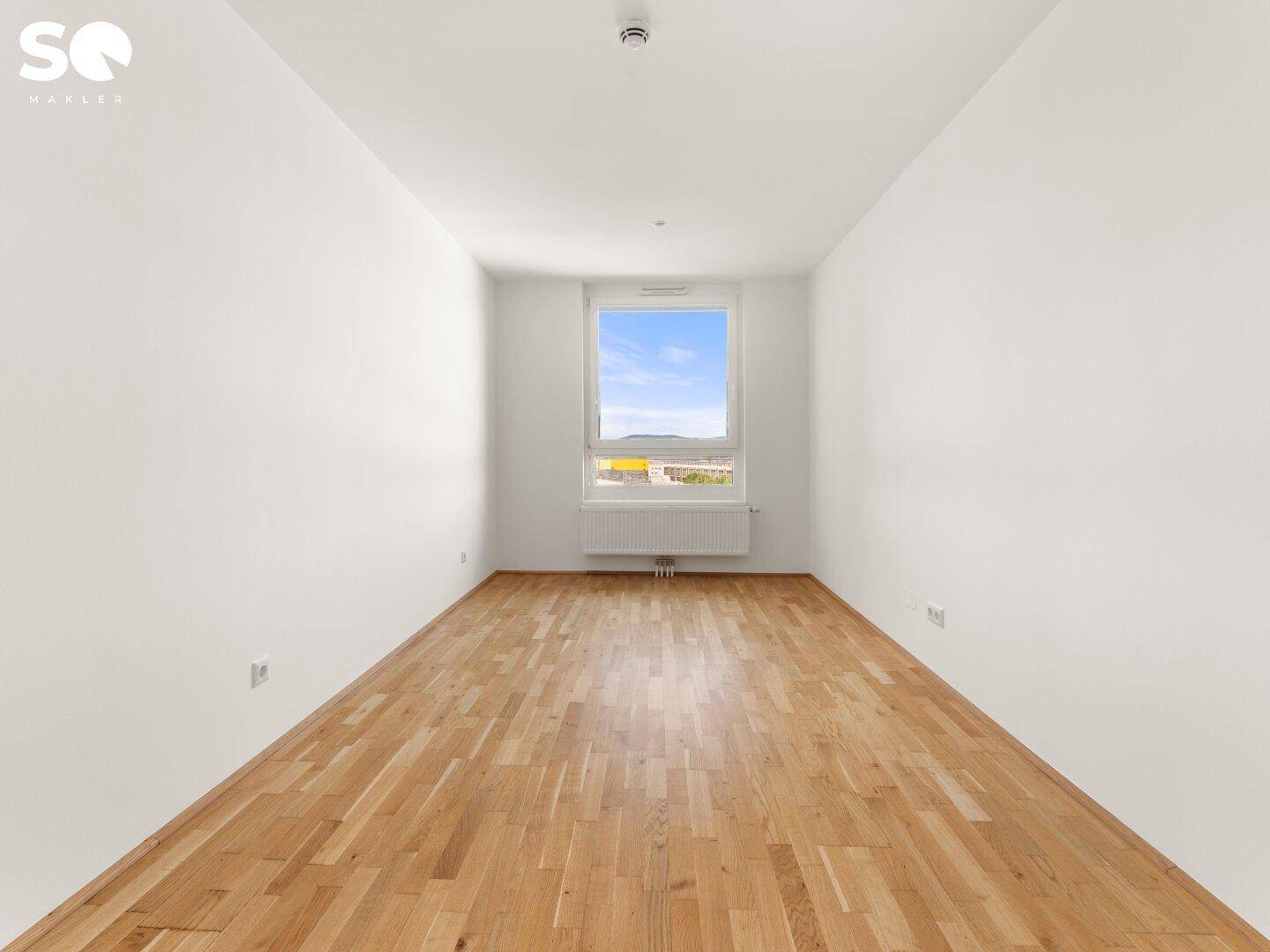 Leeres Zimmer mit Parkettboden und einem Fenster, das viel Tageslicht hereinlässt.