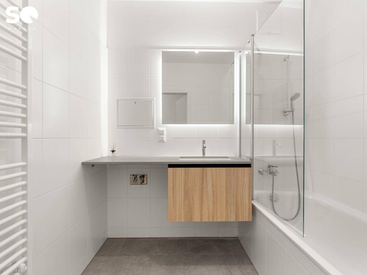 Modernes Badezimmer mit weißen Fliesen, Waschtisch, beleuchtetem Spiegel und Badewanne mit Duschabtrennung.