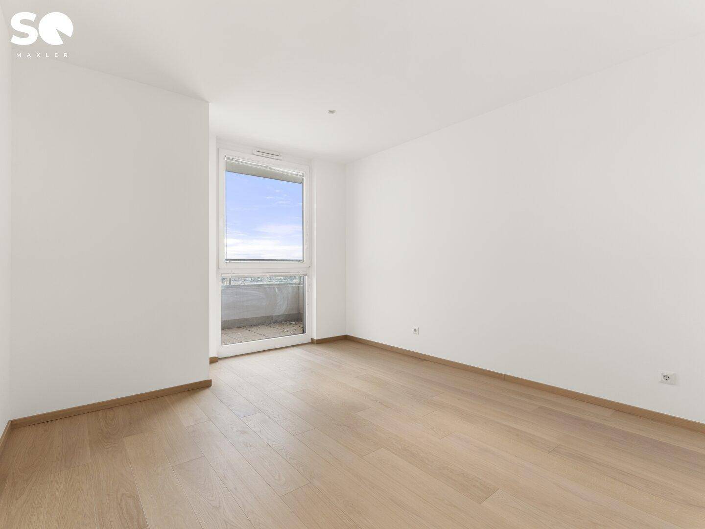 Helles, leeres Zimmer mit Parkettboden und großem Fenster, das Zugang zu einem Balkon bietet.