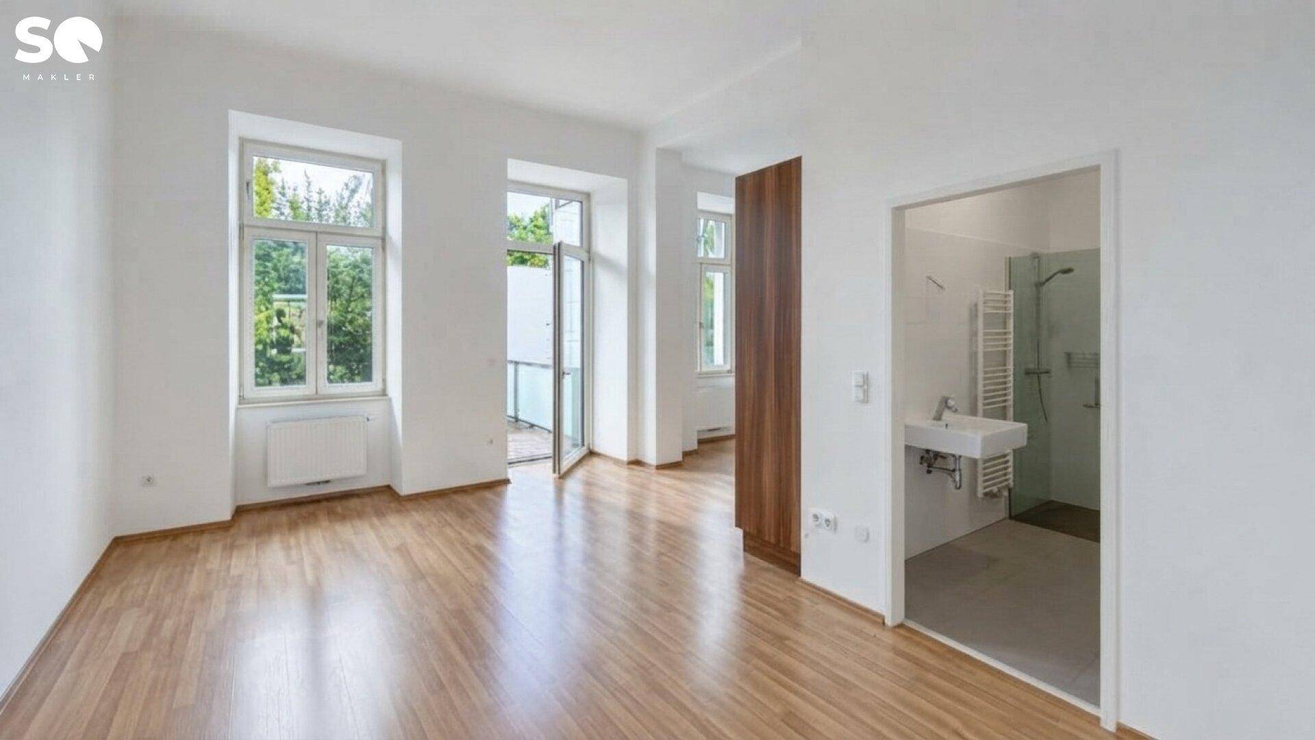 Geräumiger Wohnraum mit großen Fenstern, Holzboden und direktem Zugang zu einem kleinen Badezimmer.