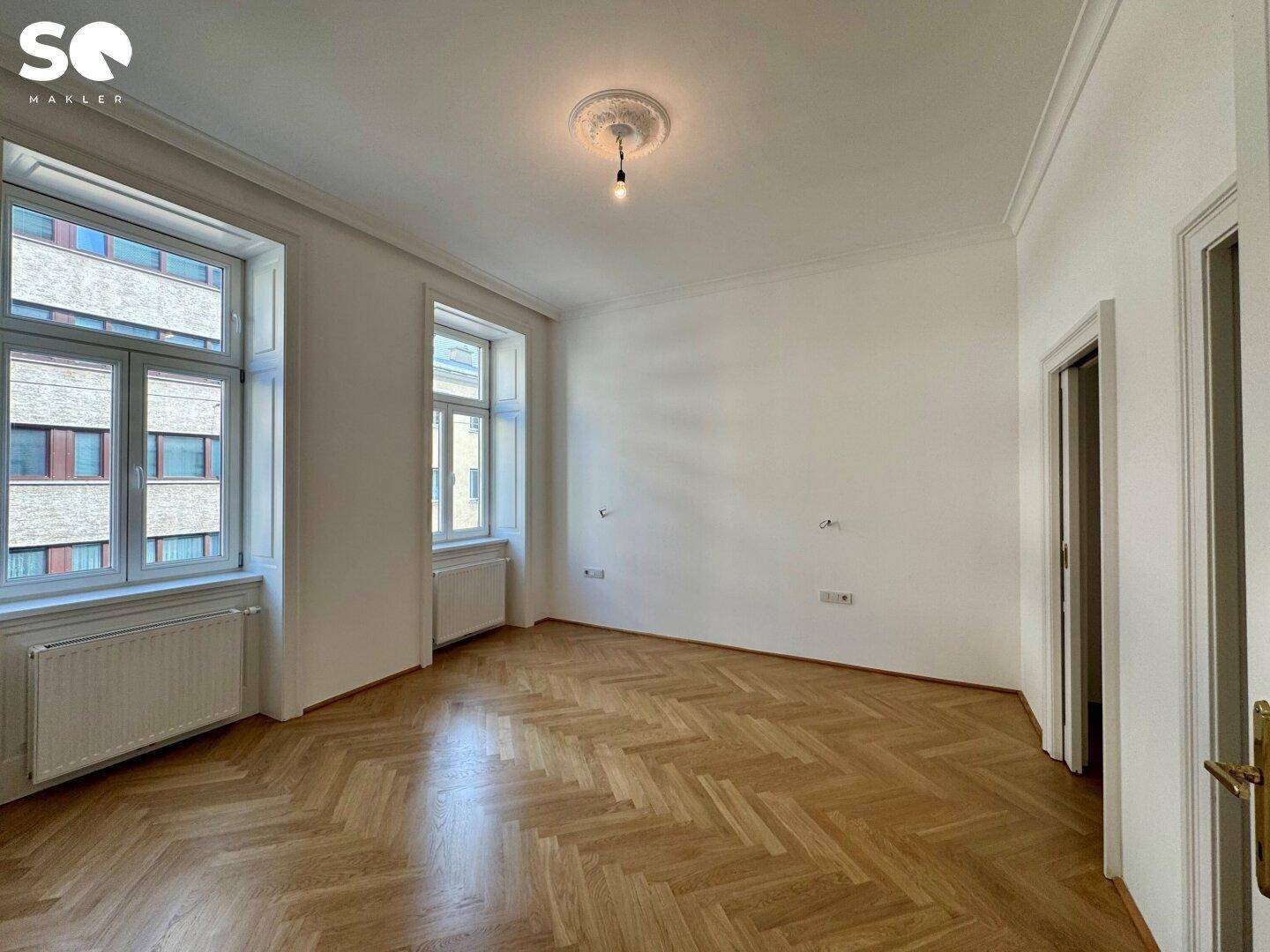 Geräumiges Zimmer mit klassischem Fischgrätparkett und zwei großen Fenstern für viel Tageslicht.
