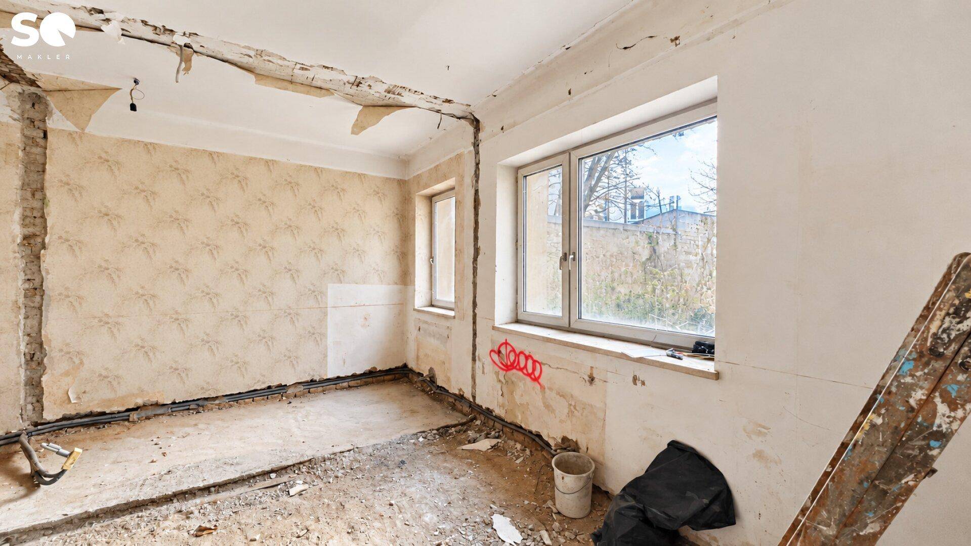 Unfertiger Innenraum mit zwei Fenstern, Betonboden und Spuren früherer Tapeten.