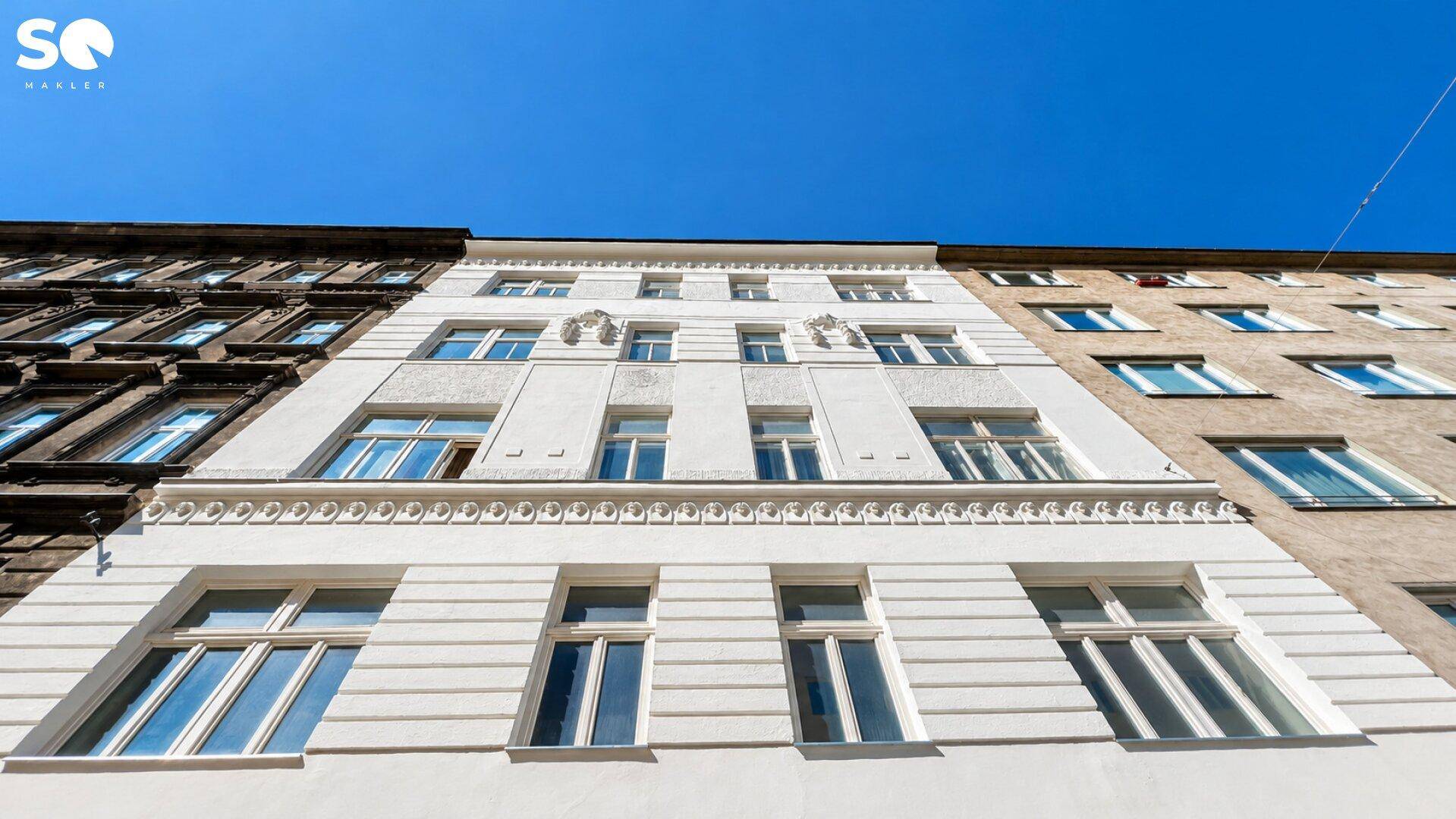 Ansicht einer weißen Gebäudefassade mit vielen Fenstern und dekorativen Elementen unter blauem Himmel.