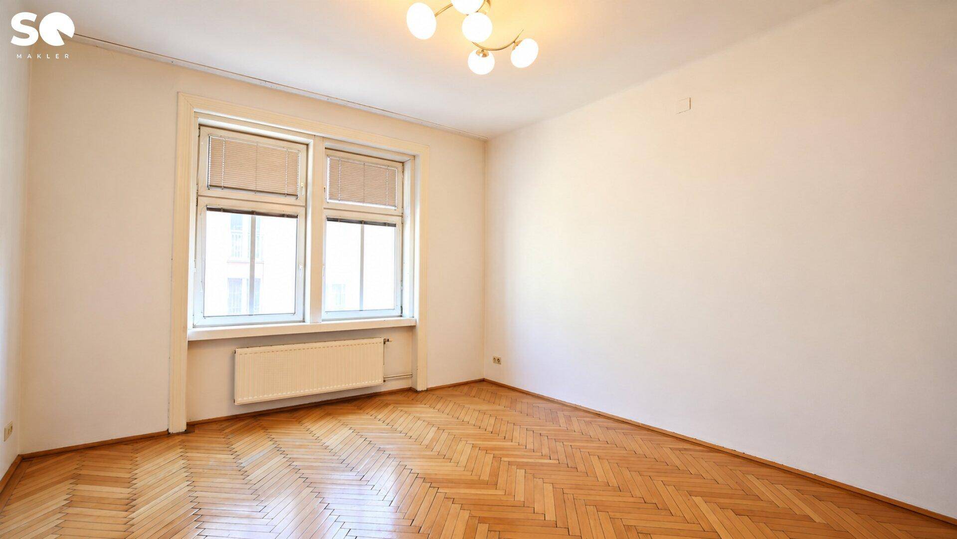 Geräumiges Wohnzimmer mit Fischgrätparkett, großen Fenstern und einem Heizkörper unter dem Fenster.