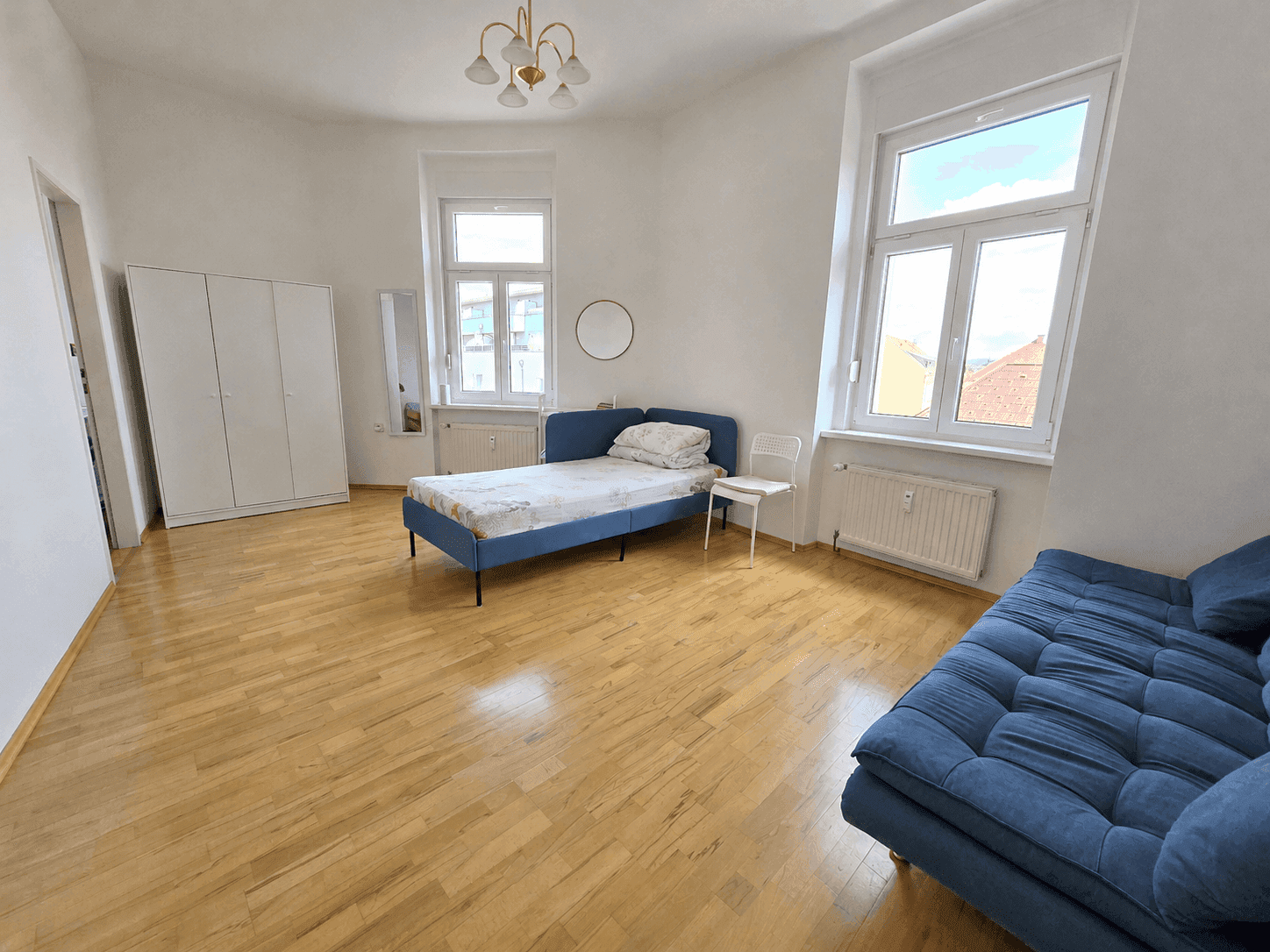 Geräumiges Schlafzimmer mit Holzboden, blauer Schlafcouch, weißem Kleiderschrank und viel Tageslicht.