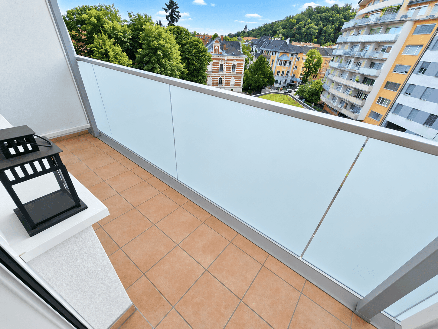 Moderner Balkon mit Fliesenboden und Glasgeländer, bietet einen Blick auf die Stadt und viel Grün.