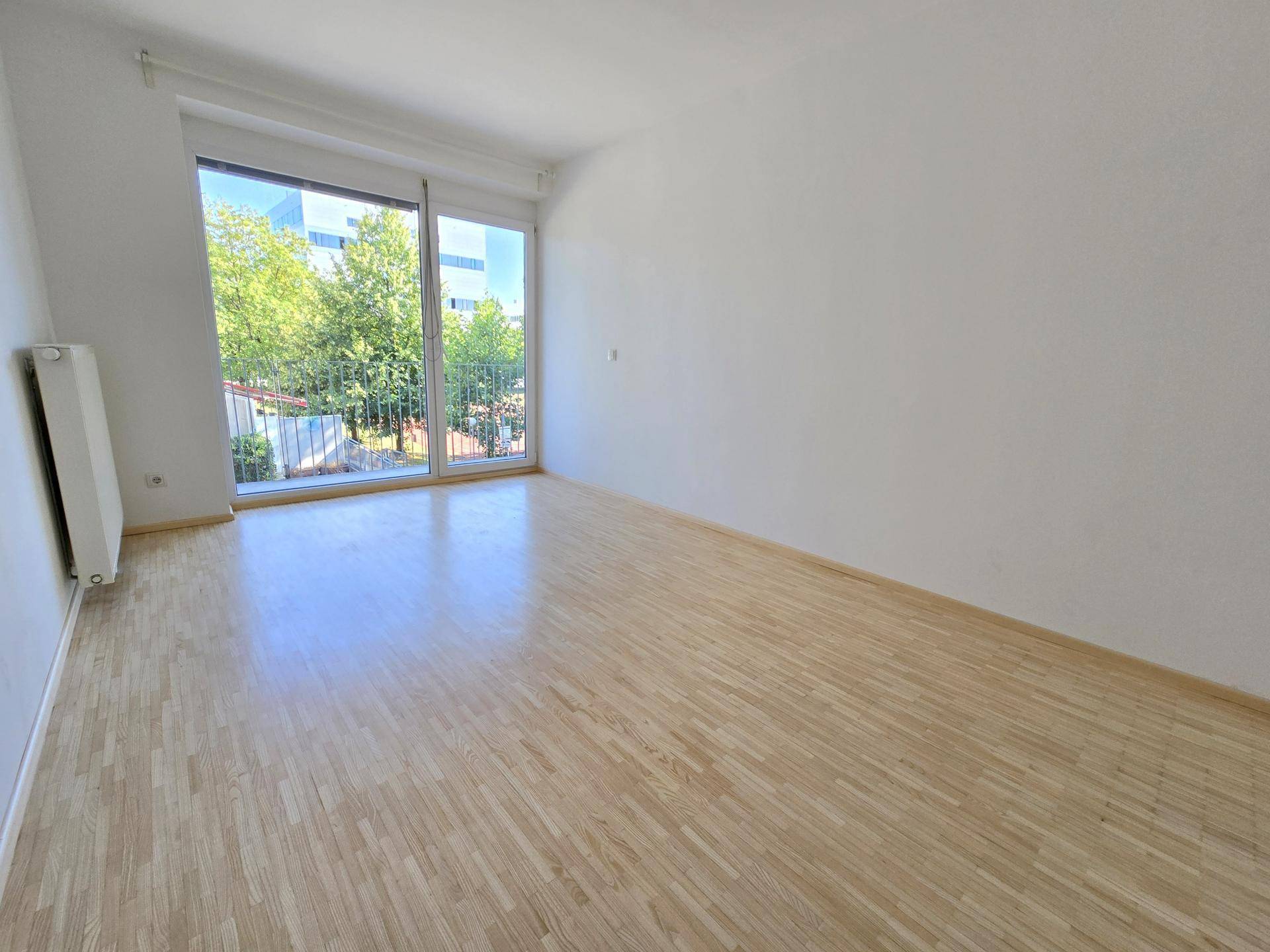Heller, leerer Raum mit Laminatboden, weißen Wänden und großem Fenster mit Blick ins Grüne.