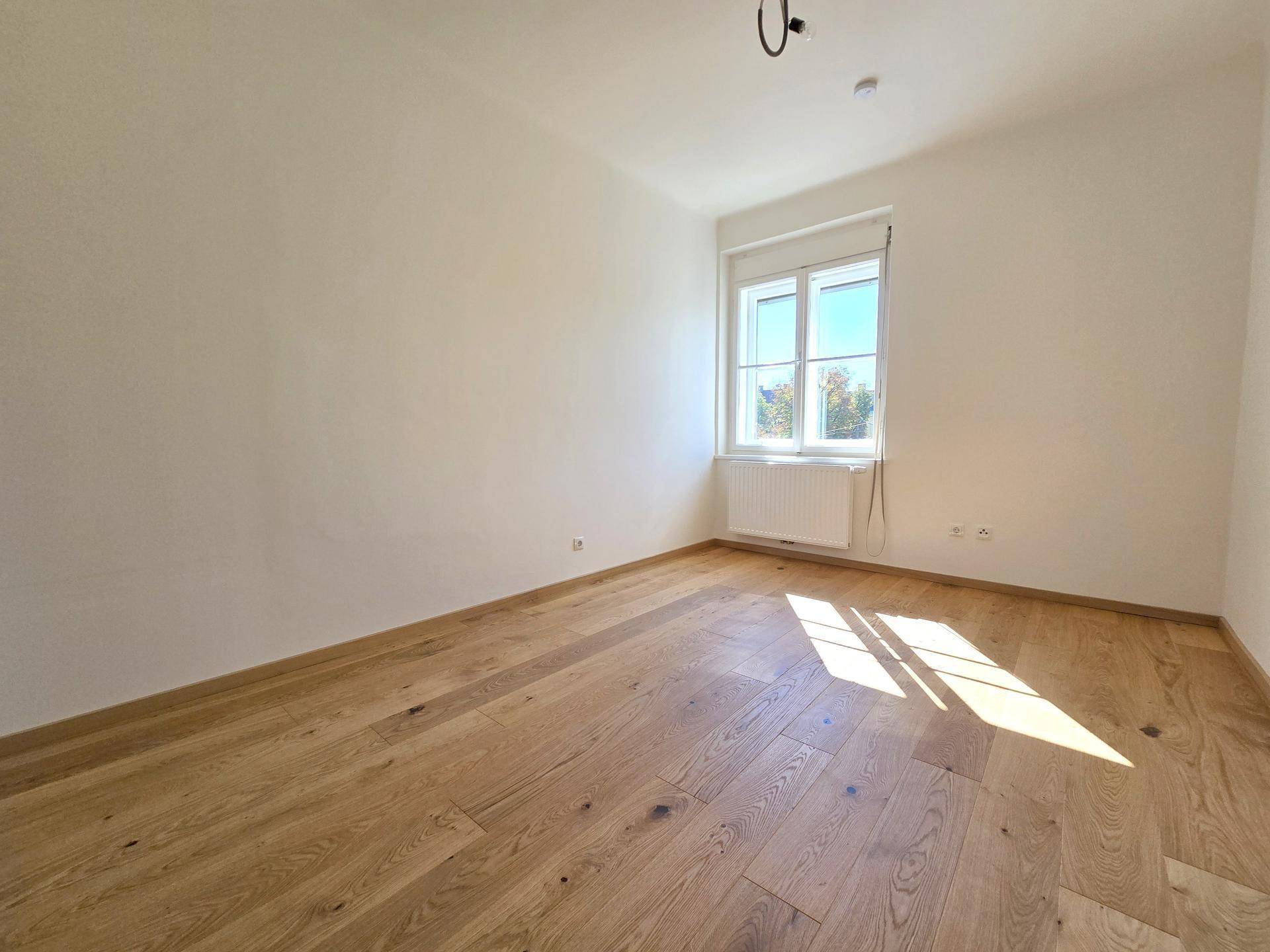 Lichtdurchfluteter Raum mit Holzboden und einem Fenster, das einen Blick ins Grüne bietet.