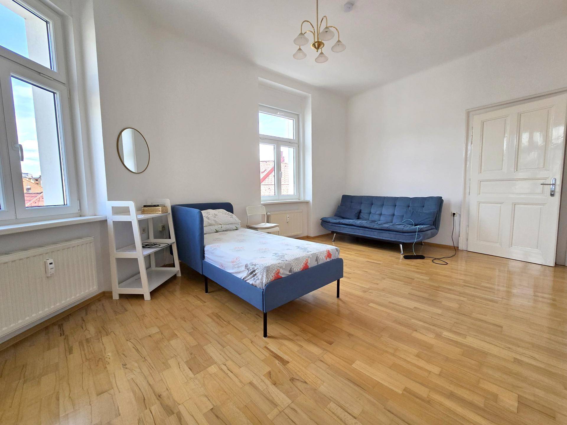 Helles Schlafzimmer mit Holzboden, einem blauen Bett, Sofa und großen Fenstern.
