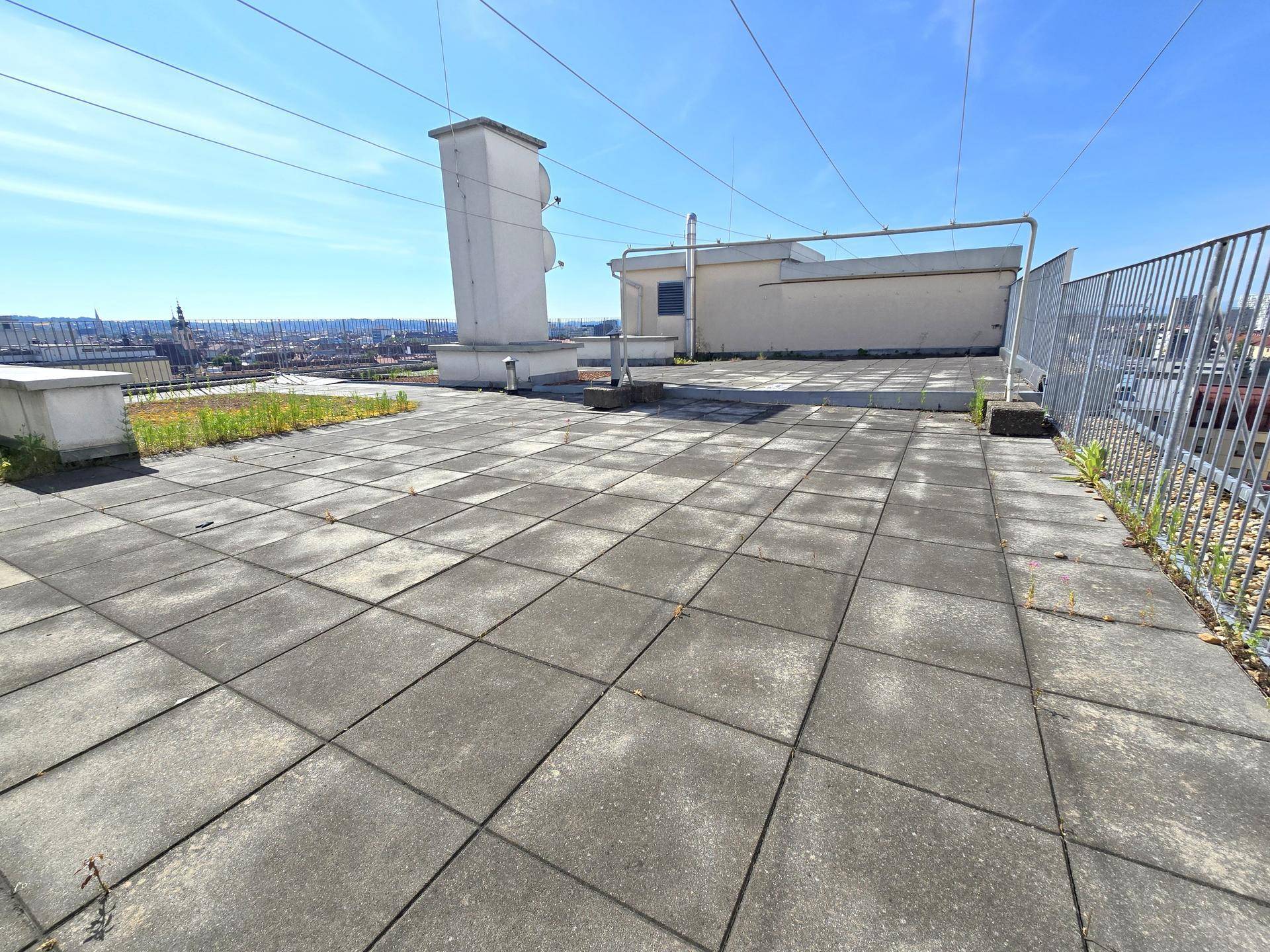 Große Dachterrasse mit Betonplatten und weitem Blick über die Stadt unter blauem Himmel.