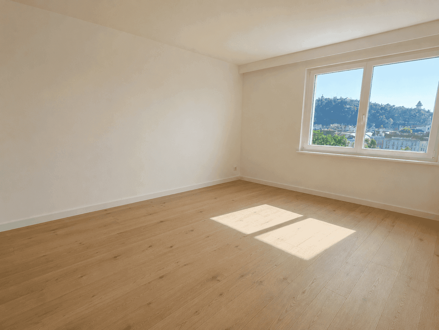 Heller, leerer Raum mit Holzfußboden und großem Fenster mit Blick auf die Stadt und Hügel.