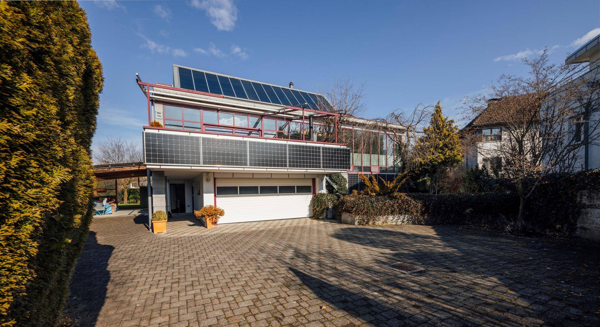 Fassade des Hauses mit integrierter Garage, gepflasterter Auffahrt und Solarpaneelen an der Wand.