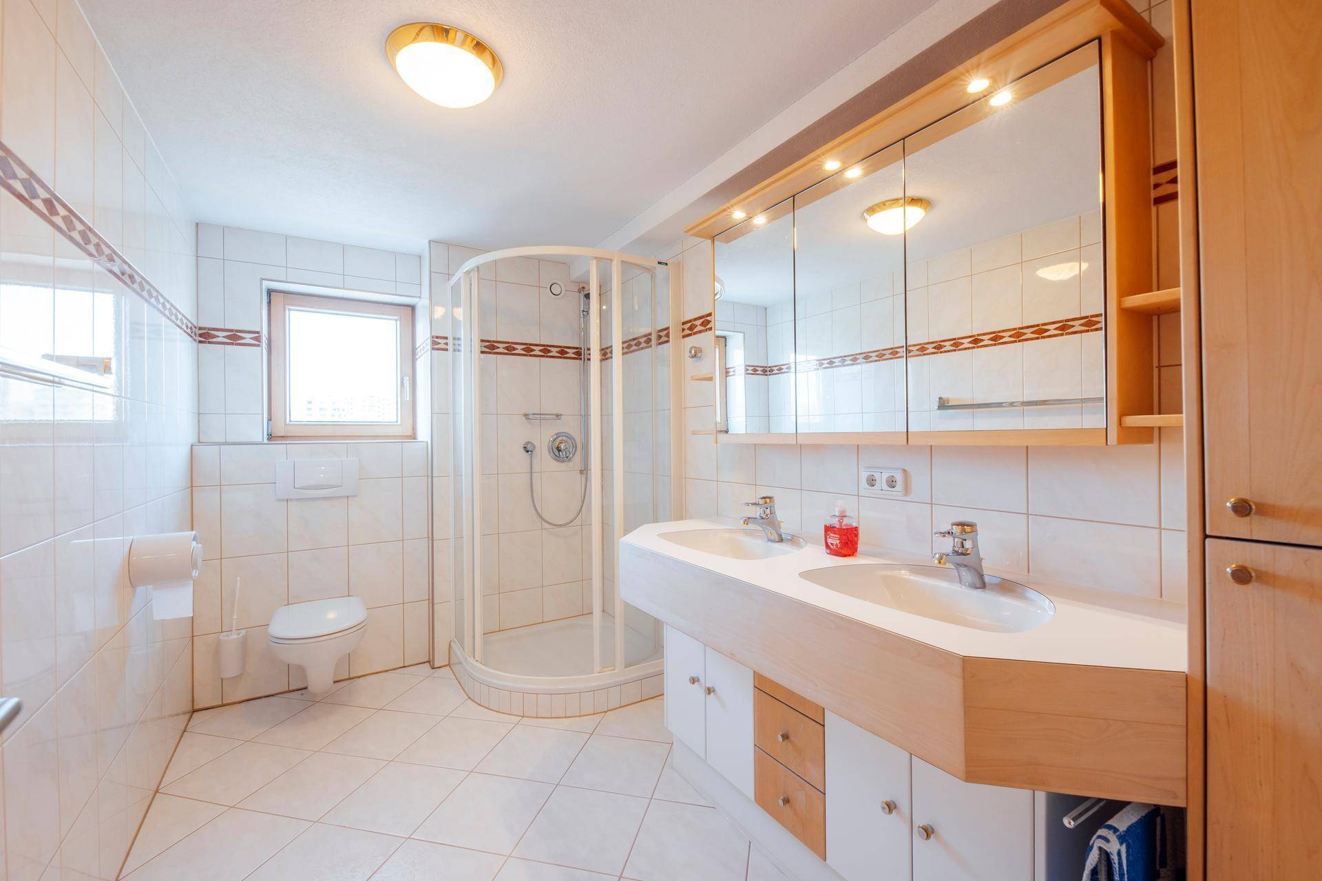 Hell gefliestes Badezimmer mit Dusche, Doppelwaschbecken, Spiegelschrank und einem Fenster.
