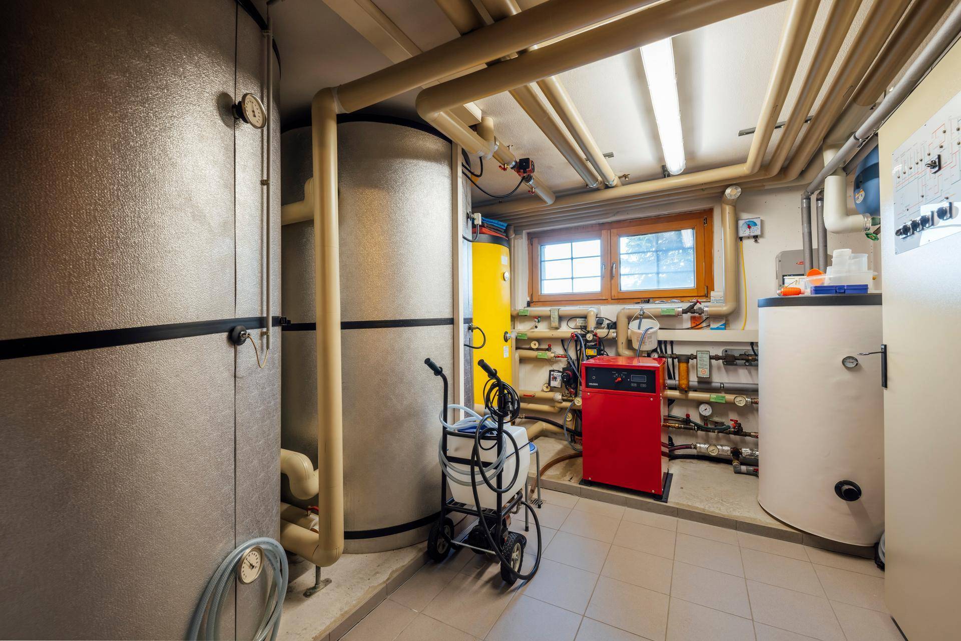 Funktionaler Technikraum im Keller mit Heizungsanlagen, Warmwasserspeichern und einem Fenster.