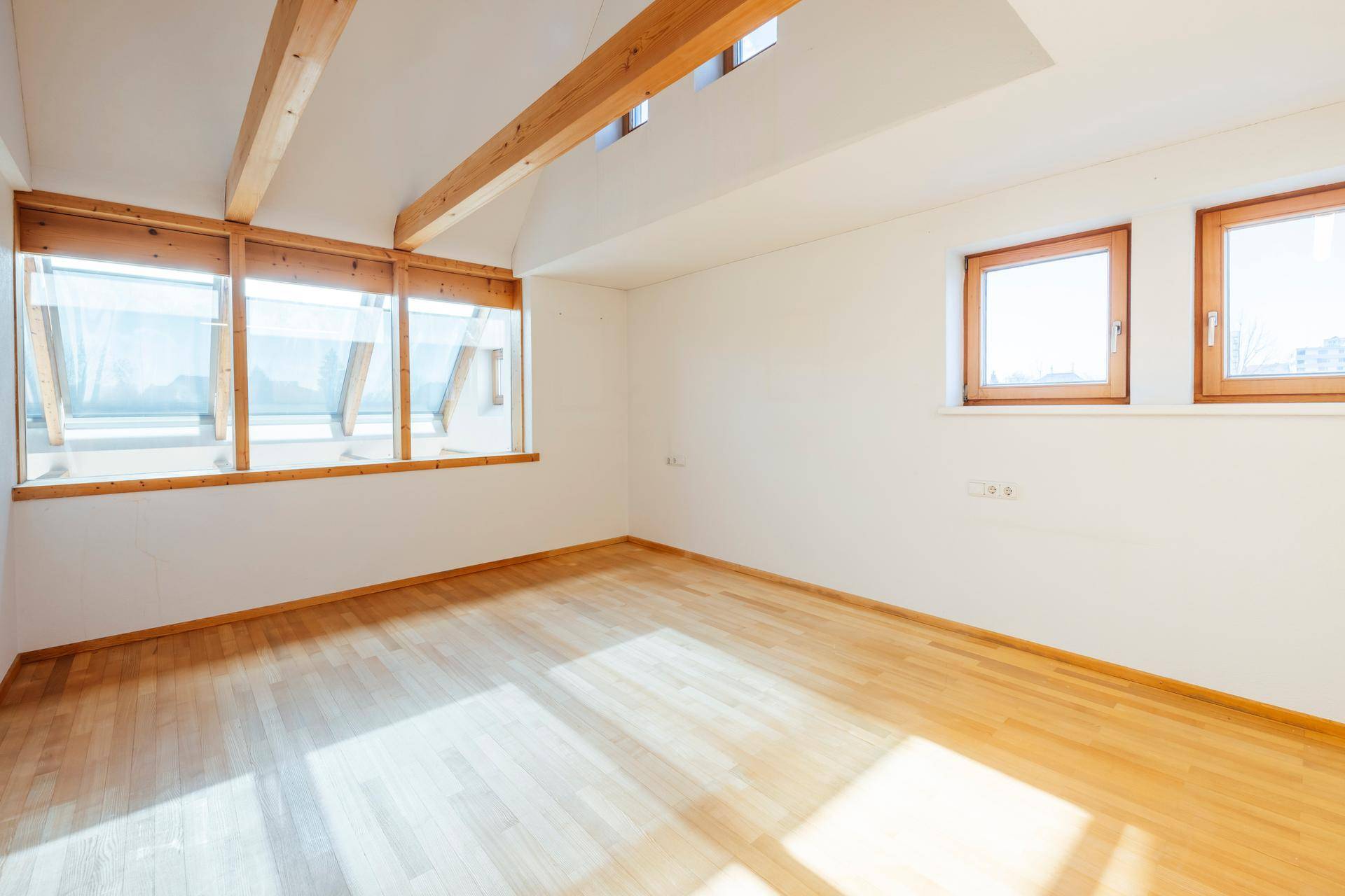 Helles Zimmer mit Holzboden, Dachschrägen und großen Fenstern, die viel Tageslicht bieten.