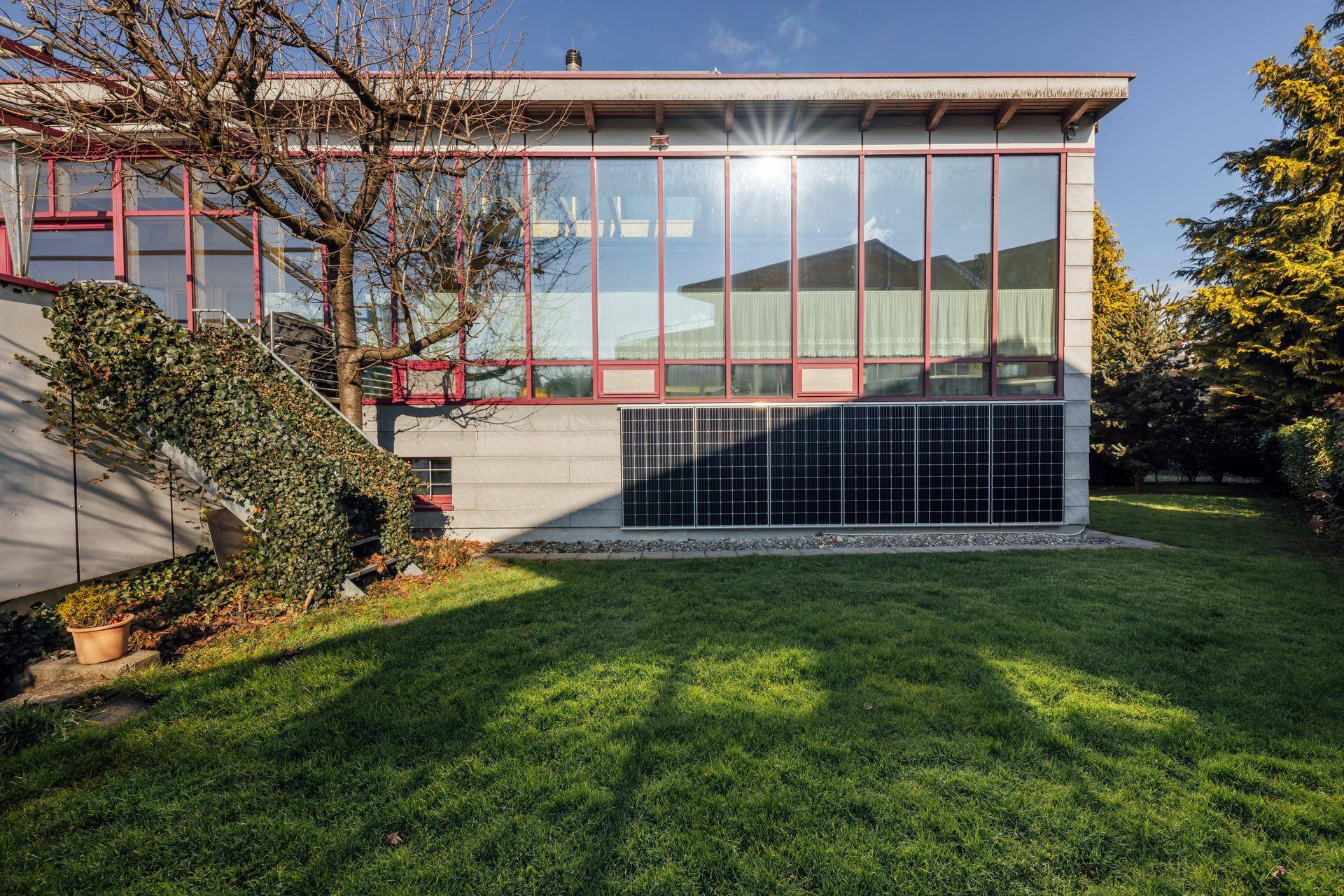 Moderne Hausfassade mit großen Fensterfronten, Solarpaneelen und einem gepflegten Rasenbereich.