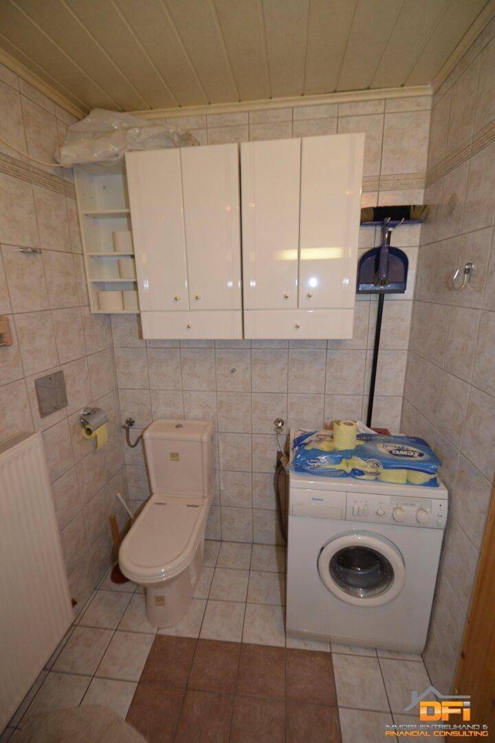 WC-Bereich mit Toilette, Waschmaschine und Hängeschränken auf Fliesenboden.