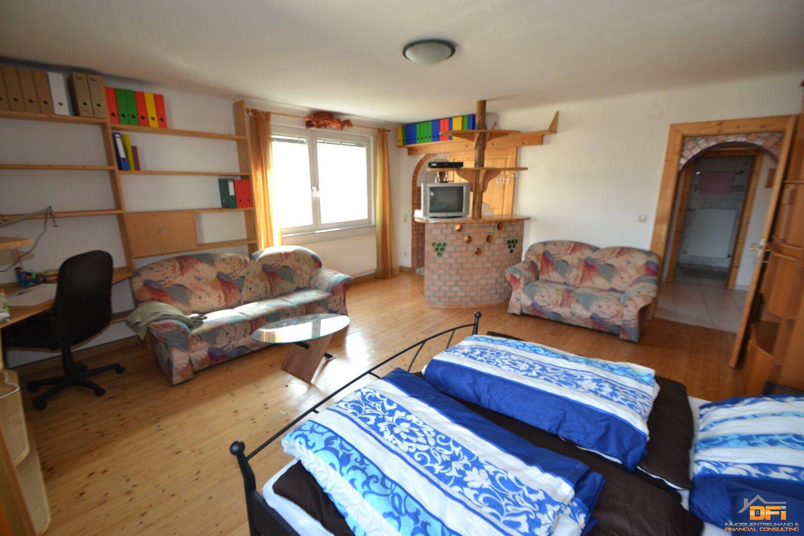 Geräumiges Schlafzimmer mit Holzdielenboden, zwei Fenstern, Sofa und Regalen.