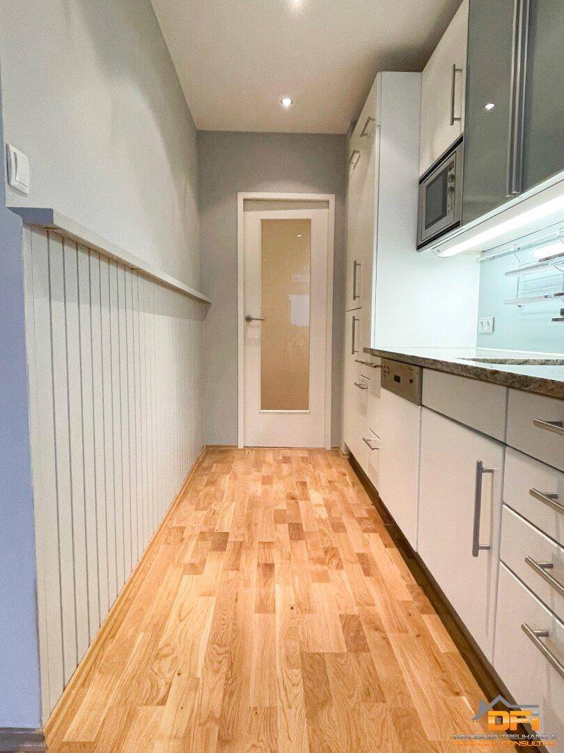 Flur mit Holzvertäfelung an einer Wand und hellem Parkettboden, führt zur Küche.