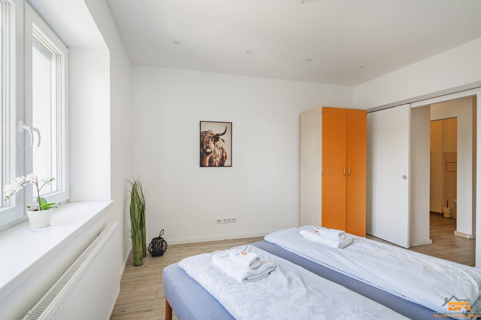 Schlafzimmer mit Doppelbett, Fenster und einem orangefarbenen Kleiderschrank mit Schiebetüren.