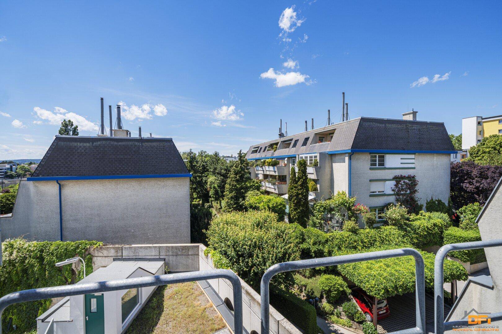 Blick vom Balkon auf die grüne Umgebung und umliegende Wohngebäude unter blauem Himmel.