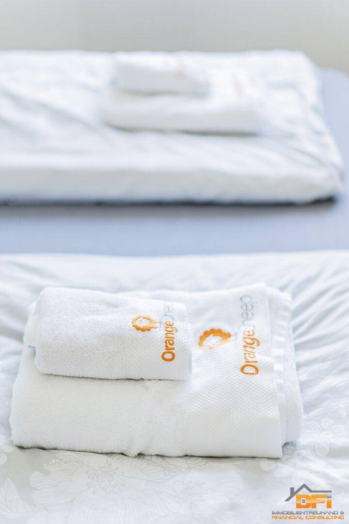 Detailansicht der weißen Handtücher mit dem orangefarbenen Logo auf dem Bett.
