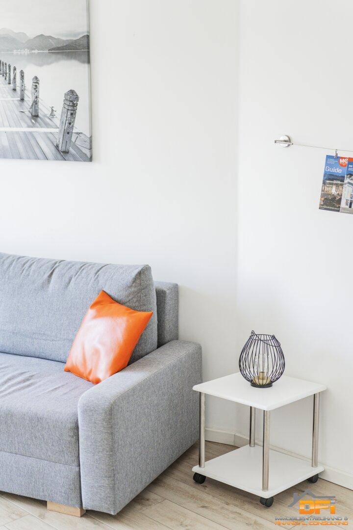 Detailansicht des Wohnbereichs mit grauem Sofa, orangefarbenem Kissen und Beistelltisch.