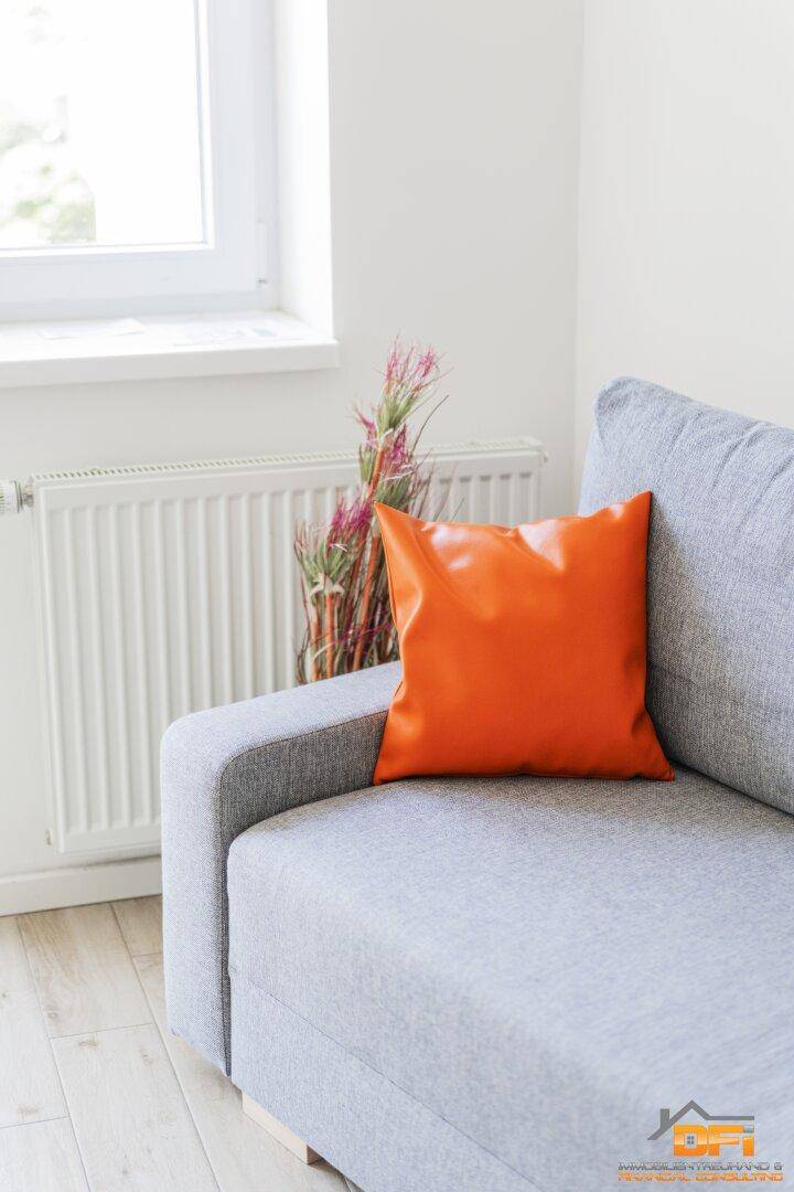 Detailansicht des Sofas mit orangefarbenem Kissen und Heizkörper unter dem Fenster.