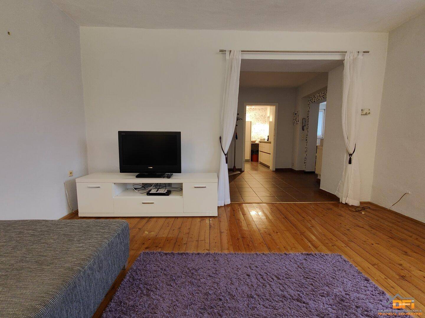 Wohnzimmer mit Holzboden, Fernseher und offenem Zugang zu einem weiteren Raum.