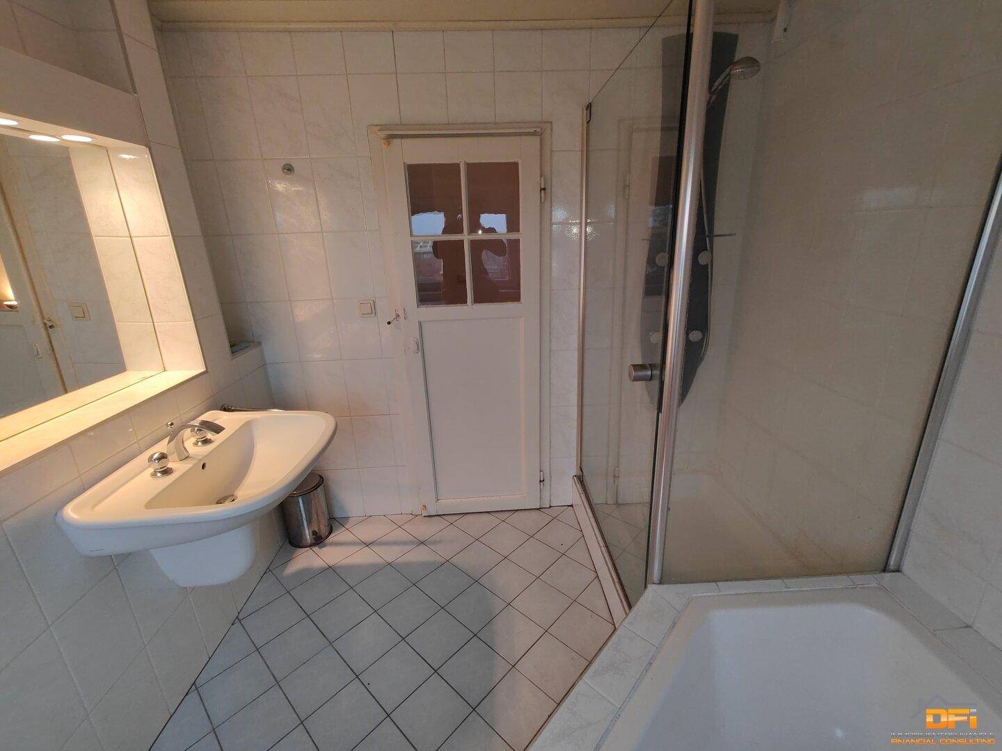 Badezimmer mit Waschbecken, Duschkabine und Tür, komplett mit weißen Fliesen gefliest.