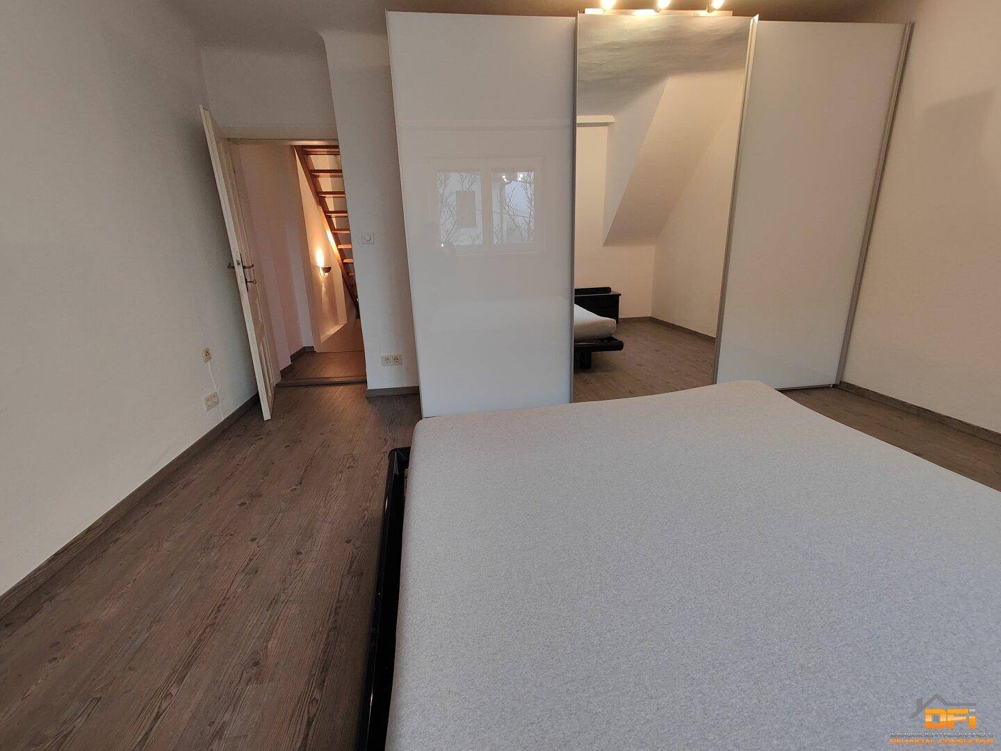 Schlafzimmer mit Doppelbett, Spiegelschrank und Zugang zum Treppenhaus auf Laminatboden.