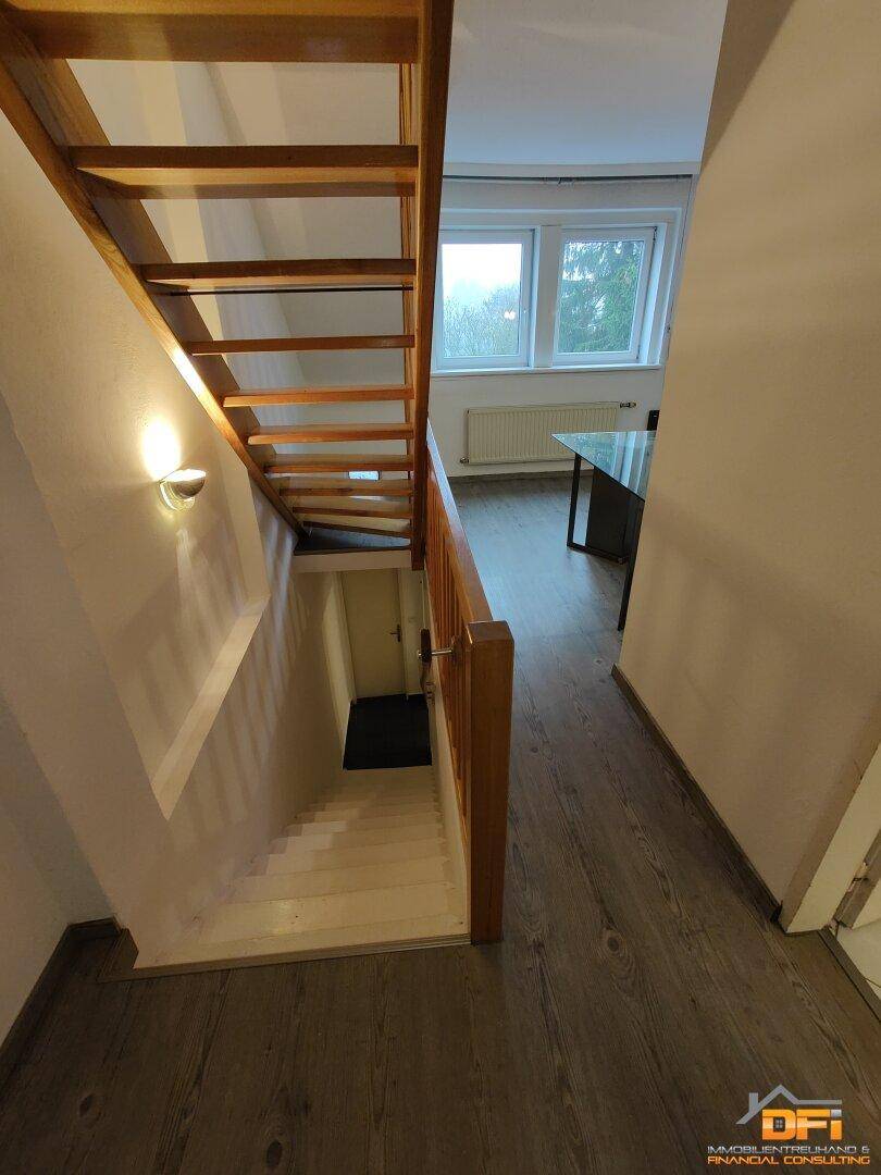 Treppenhaus mit Holzstufen und Geländer, führt zu einem hellen Raum mit Laminatboden.