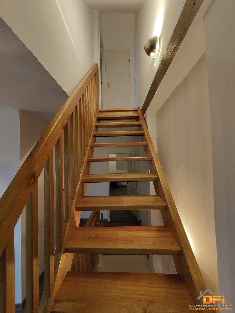 Holztreppe mit Geländer führt nach oben, beleuchtet von Wandleuchten.
