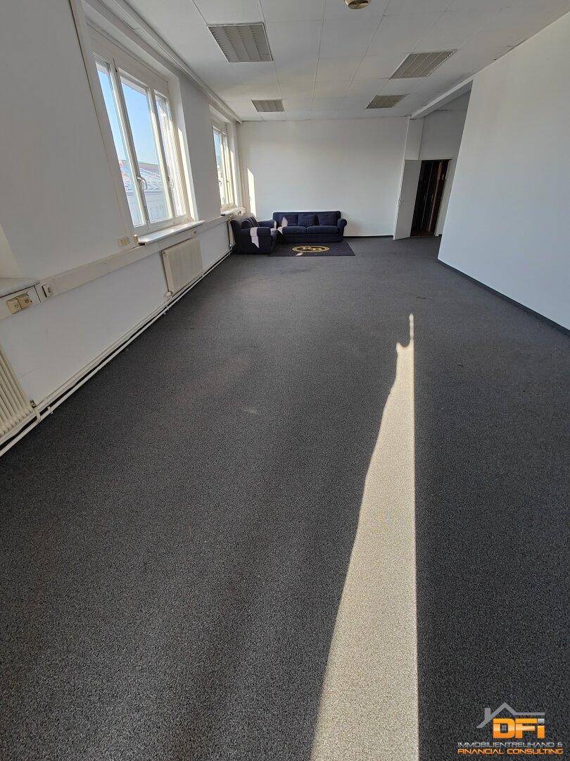 Helles Büro mit grauem Teppichboden und einer kleinen Sitzecke, ideal für flexible Arbeitsbereiche.
