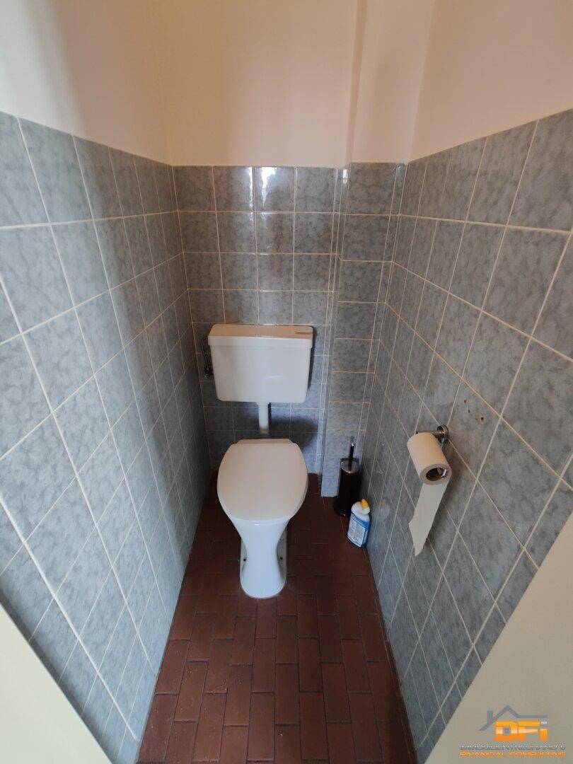 Kleines Gäste-WC mit weißer Toilette und grauen Fliesen an den Wänden sowie Terrakotta-Boden.