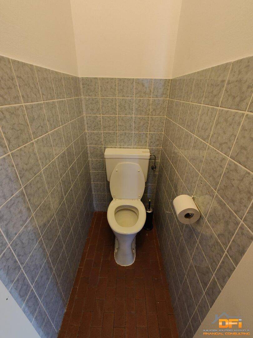 Einfaches Gäste-WC mit klassischer Toilette, umgeben von grauen Wandfliesen und Terrakotta-Boden.