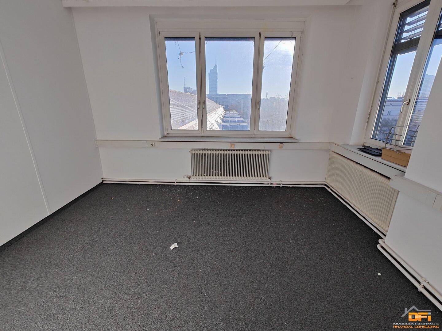 Eckbereich eines Büros mit grauem Teppichboden, weißen Wänden und Fenstern, die Tageslicht hereinlassen.