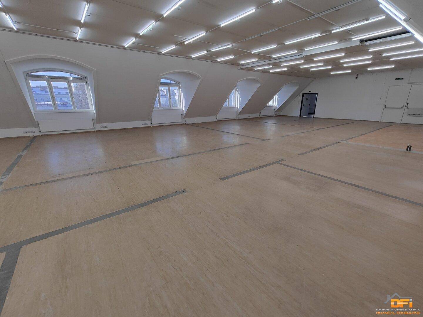 Weitläufiger Raum im Dachgeschoss mit schrägen Wänden, vielen Fenstern und heller Beleuchtung.