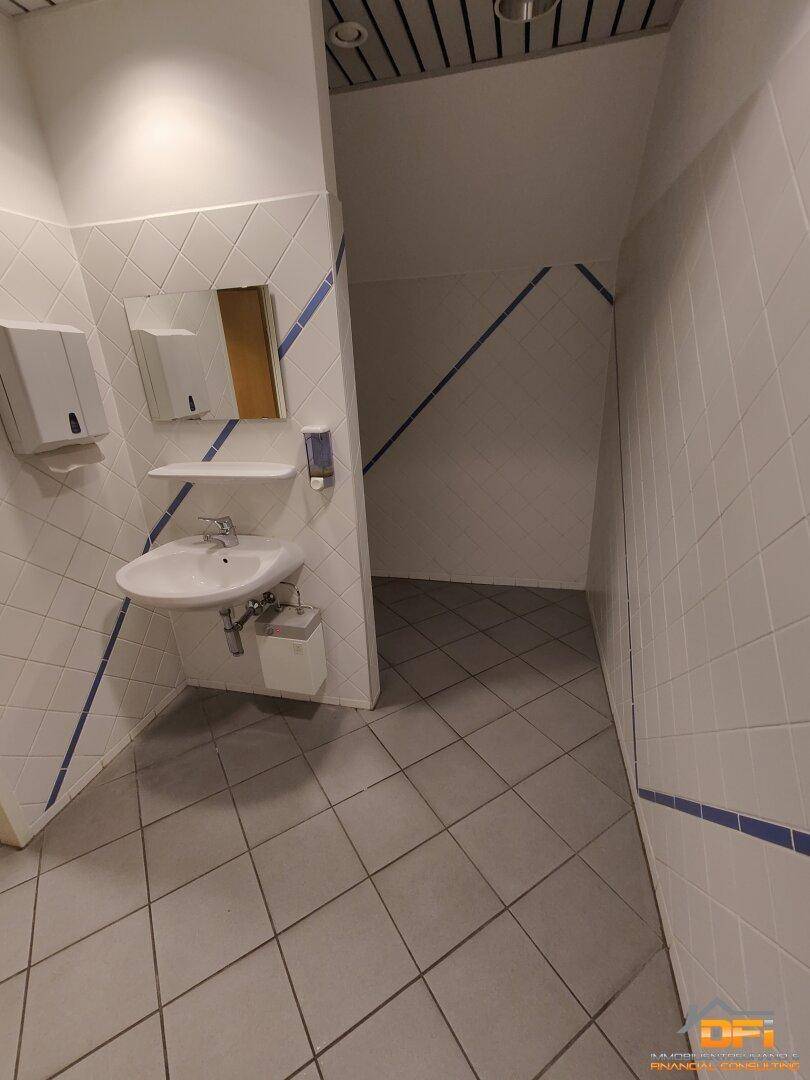 Öffentliche Toilette mit Waschbecken, Spiegel und weiß-blauen Fliesen an den Wänden.