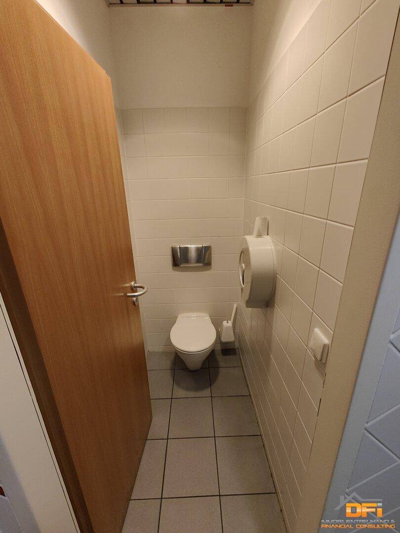 Toilettenkabine mit WC und weiß gefliesten Wänden, daneben eine Holztür.