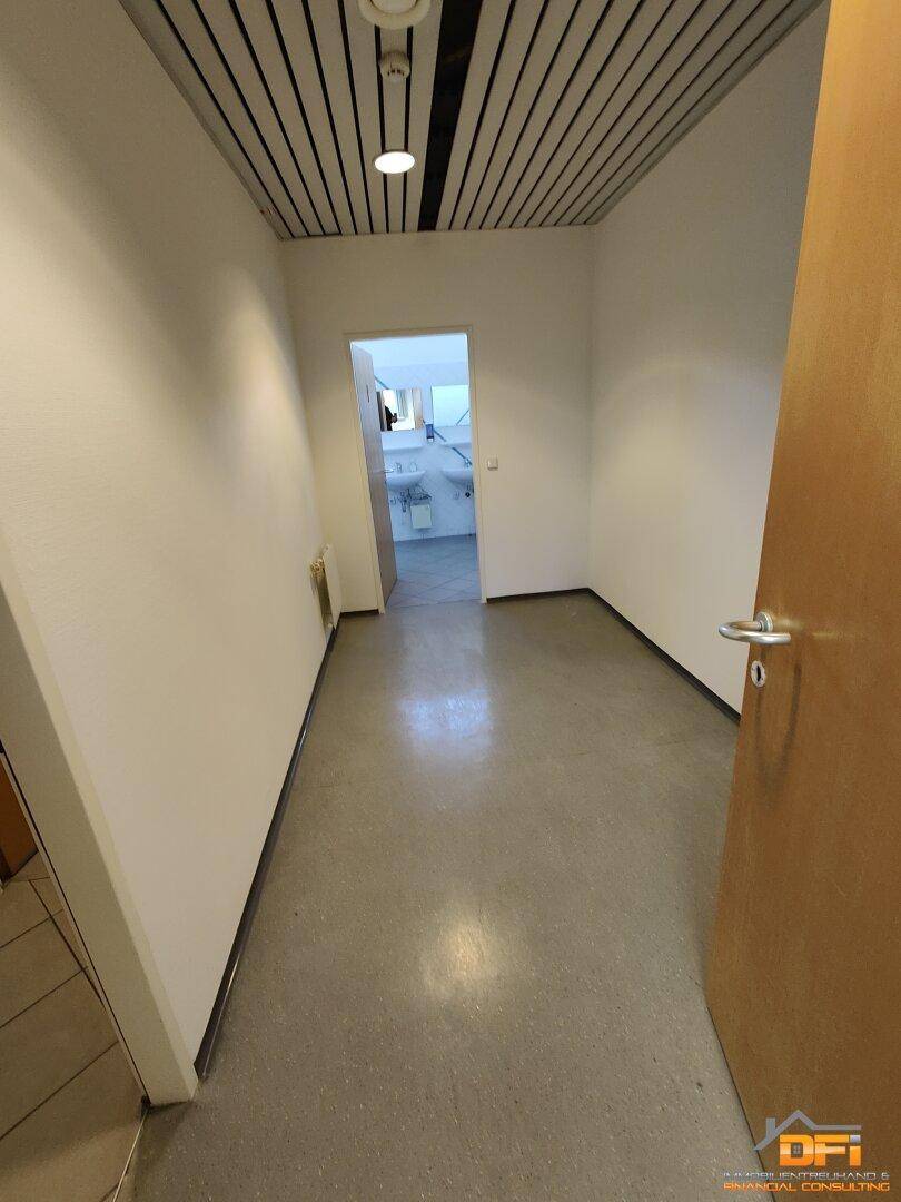 Schmaler Flur mit hellem Betonboden und einer Decke mit Holzlamellen, führt zu einem Badezimmer.