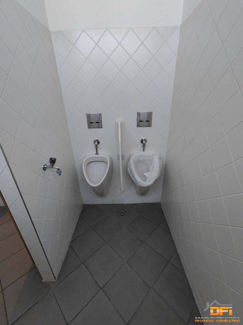 Urinalbereich in einer öffentlichen Toilette mit weißen Fliesen an Wänden und Boden.