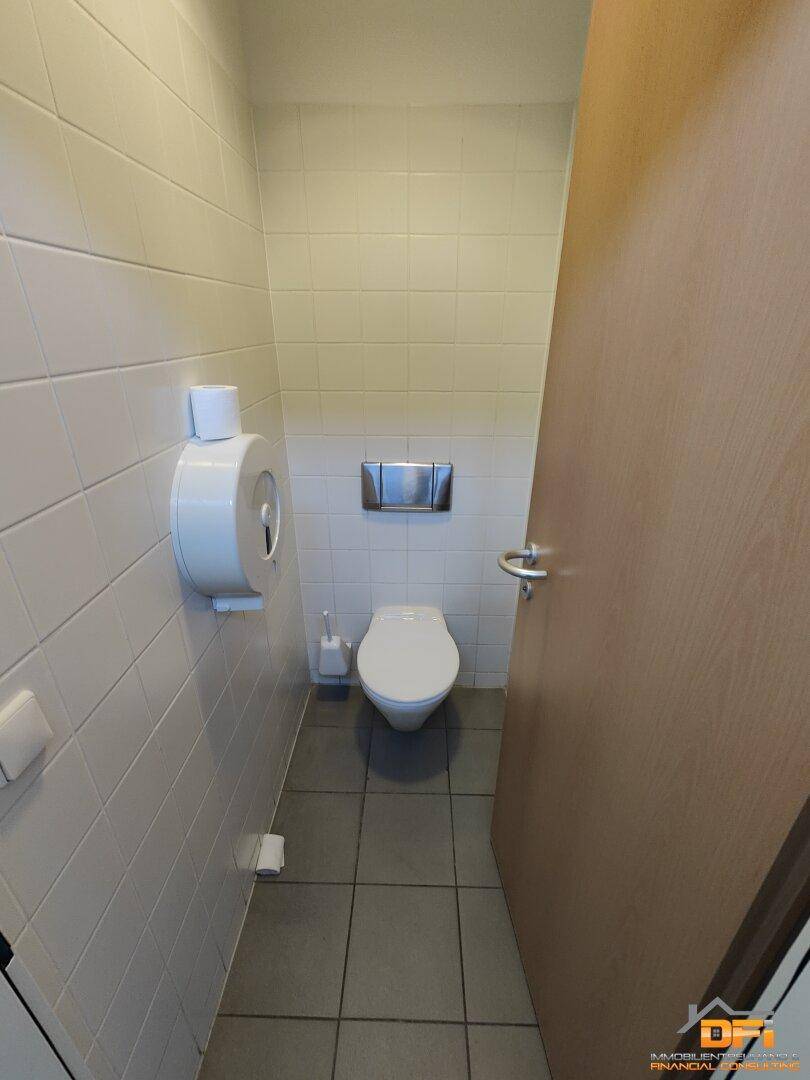 Toilettenkabine mit weißen Fliesen an den Wänden und grauem Fliesenboden, eine Holztür ist sichtbar.