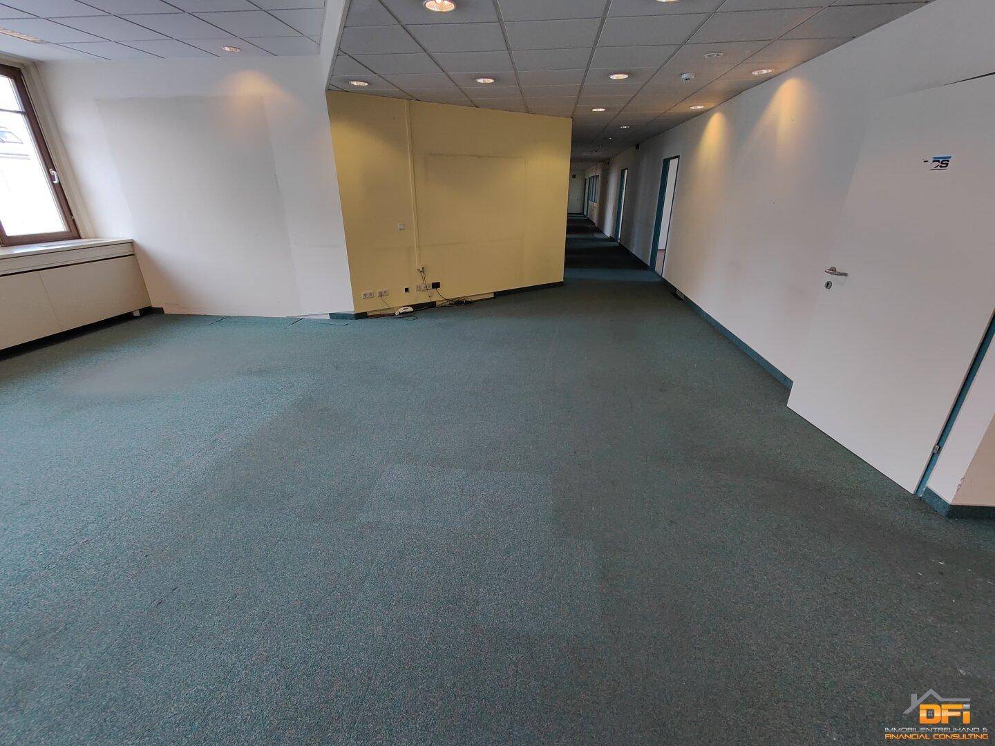 Langer Flur mit grünem Teppichboden und abgehängter Decke, der zu weiteren Büroräumen führt.