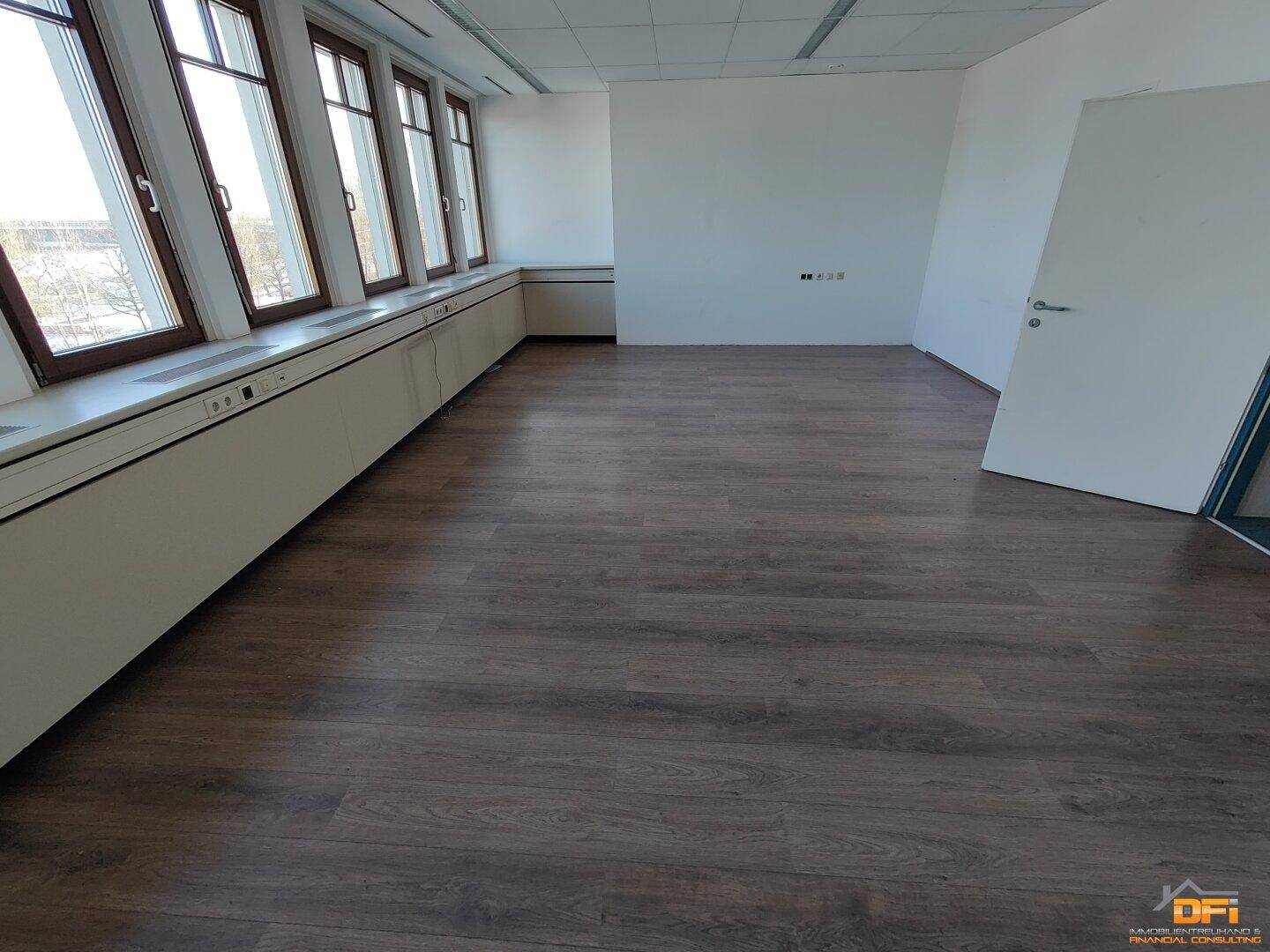 Helles Büro mit dunklem Laminatboden und einer Fensterfront, die den Raum gut belichtet und eine Aussicht bietet.
