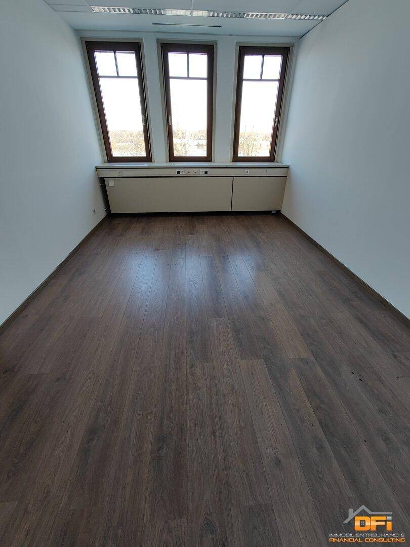 Büro mit dunklem Laminatboden und drei Fenstern, die eine weite Aussicht auf die Umgebung ermöglichen.