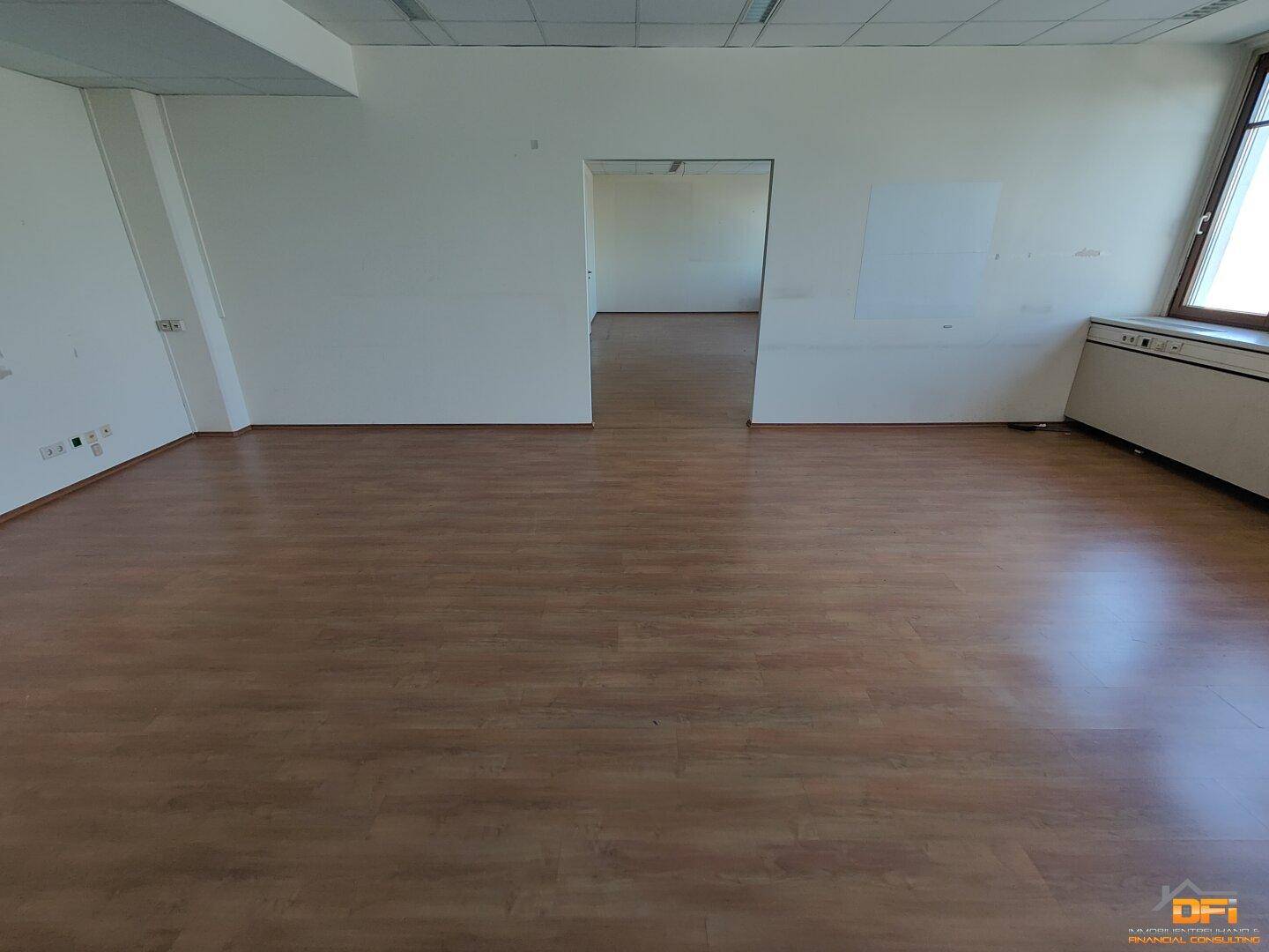 Großes, leeres Büro mit hellem Laminatboden und einem offenen Durchgang zu einem weiteren Bereich.