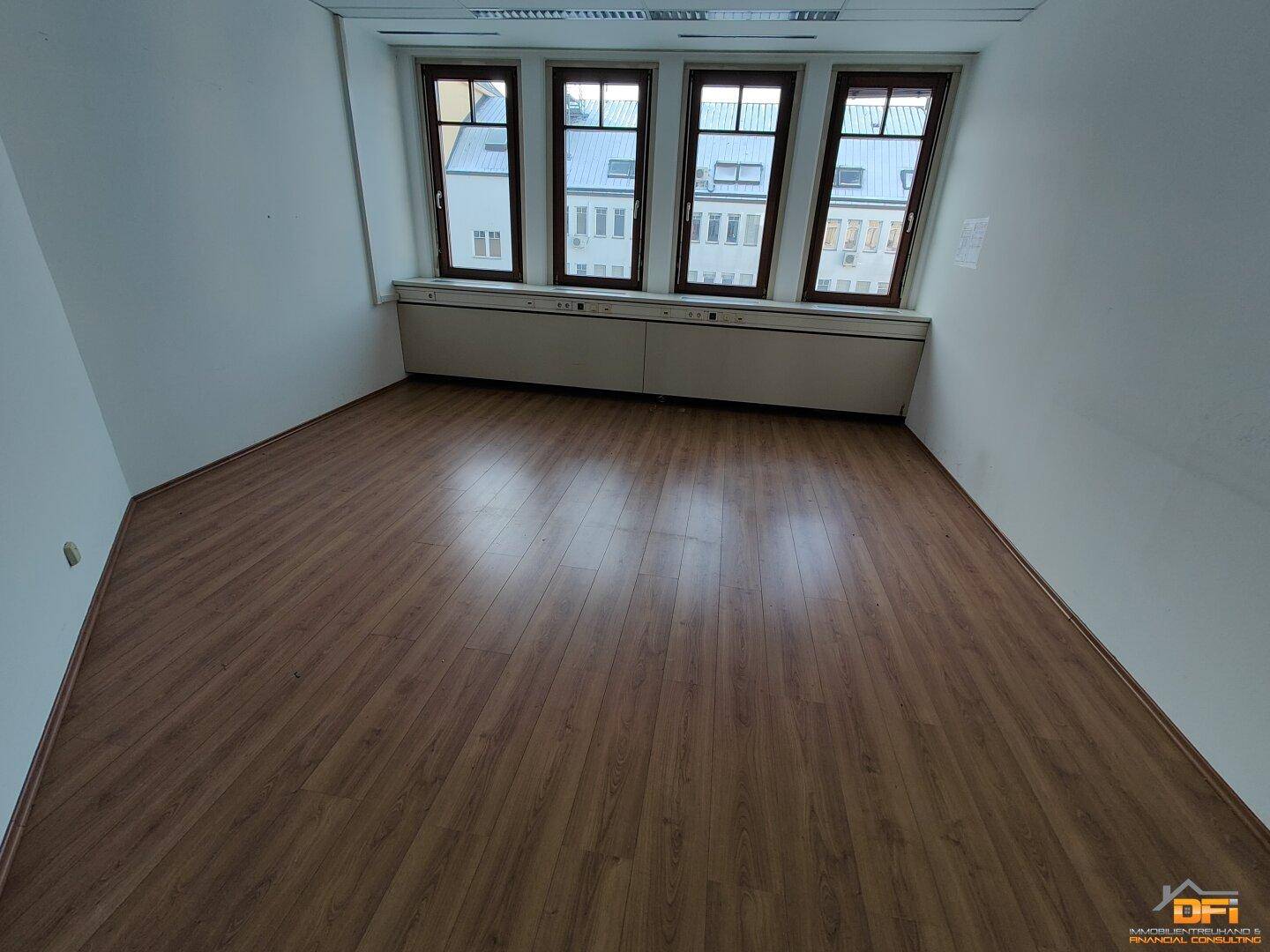 Büro mit hellem Laminatboden und vier Fenstern, die einen Blick auf die umliegenden Gebäude bieten.